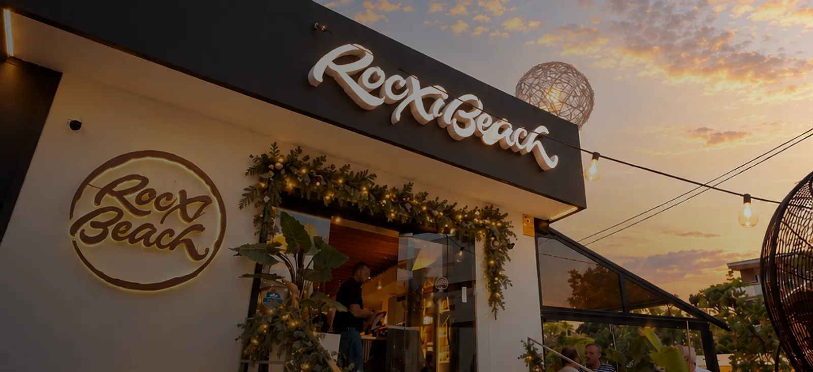 Fachada de Rocxi Beach al atardecer, con el cartel iluminado y guirnaldas